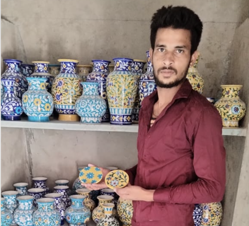 Man in maroon shirt holding two decorated ceramic coasters in front of shelves filled with colorful hand-painted vases.