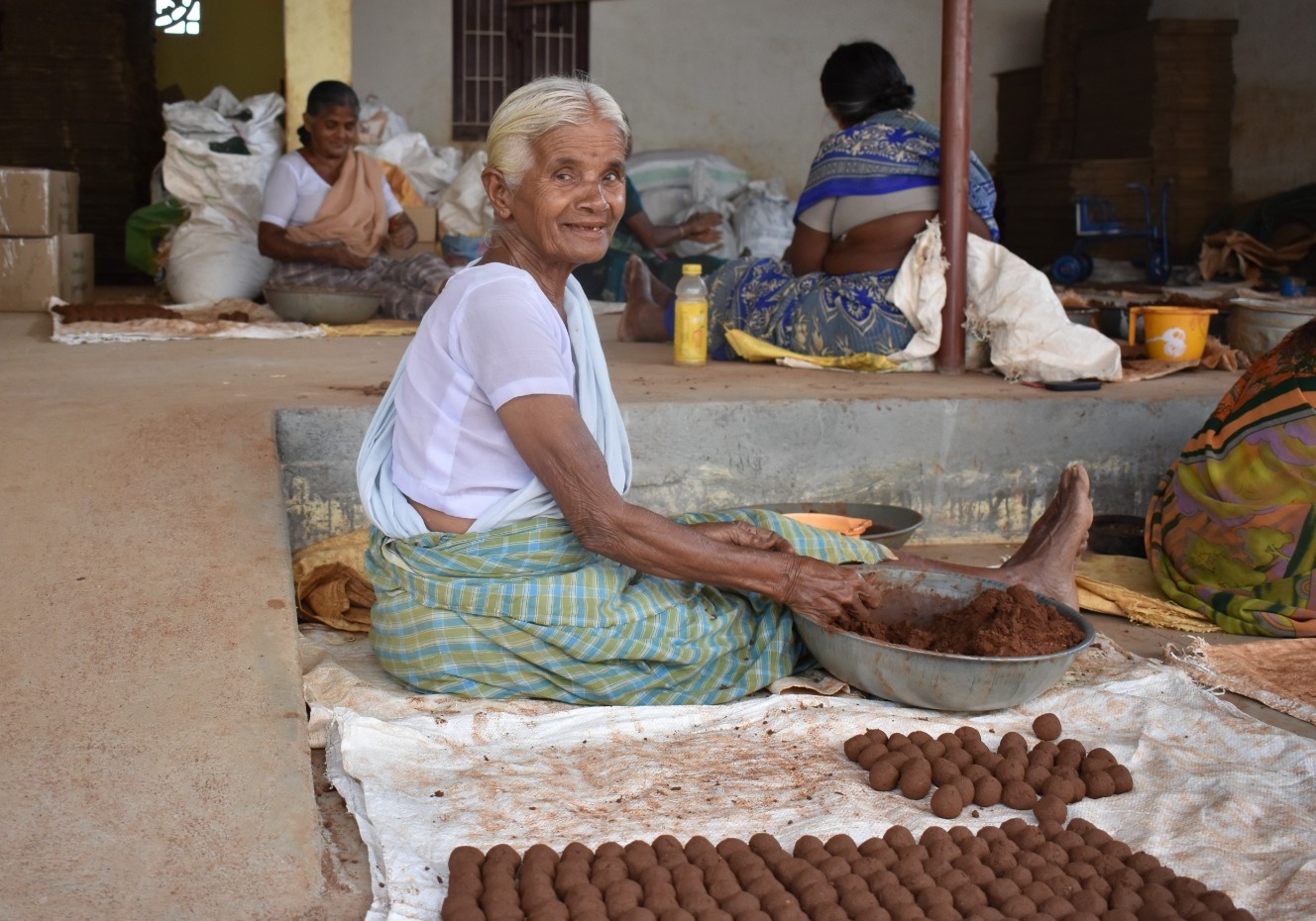 Elderly woman sitting on floor shaping balls from clay with other women working in the background.