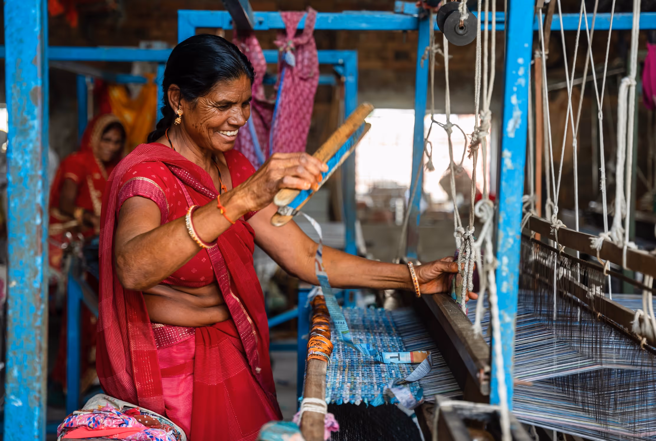 Smiling woman in a red sari weaving fabric on a traditional loom in a workshop.