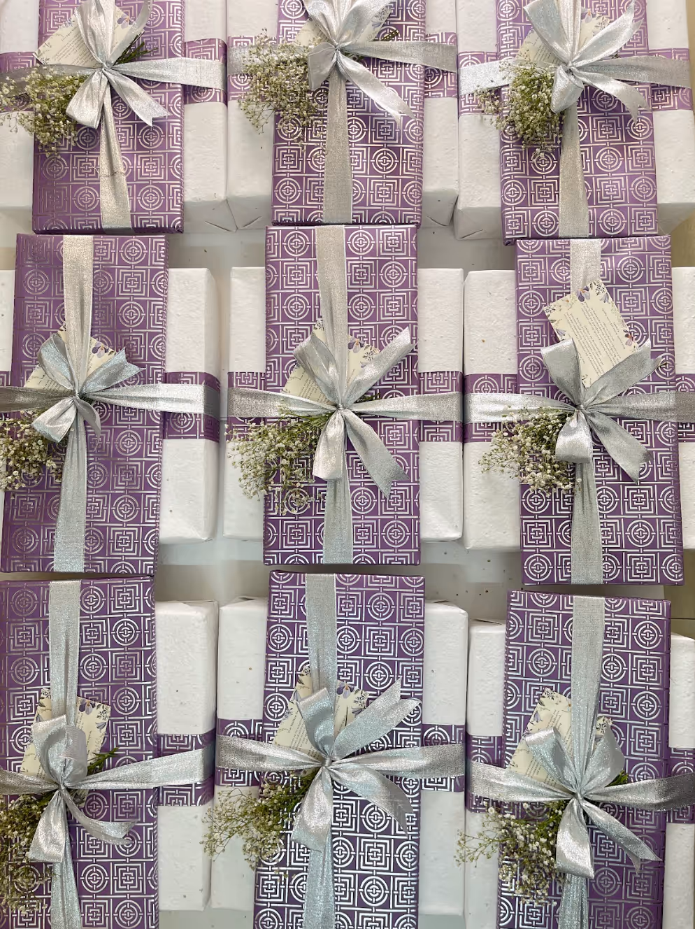 Array of rectangular gifts wrapped in white paper with purple geometric patterned bands, silver ribbons, small white flowers, and attached cards.