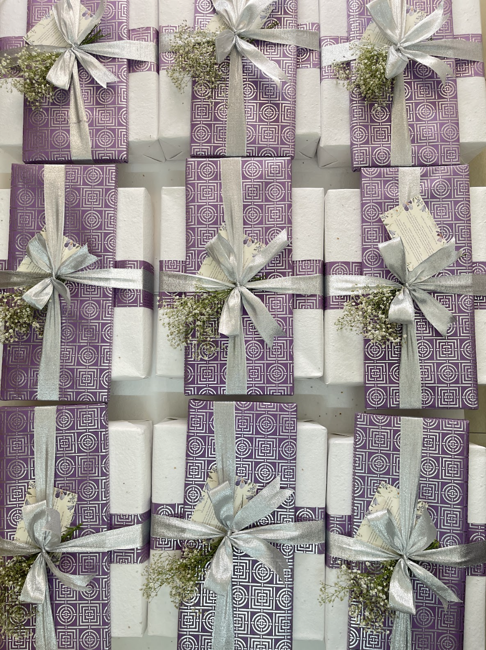 Array of rectangular gifts wrapped in white paper with purple geometric patterned bands, silver ribbons, small white flowers, and attached cards.