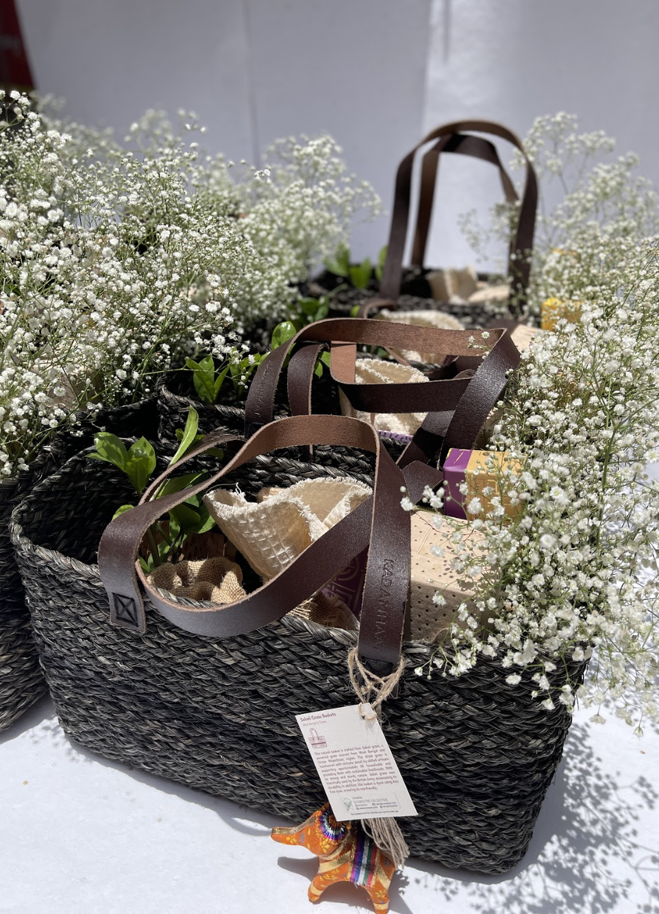 Woven dark gray basket bag with brown leather handles, filled with fabric items and decorated with white baby's breath flowers and a small colorful llama ornament.