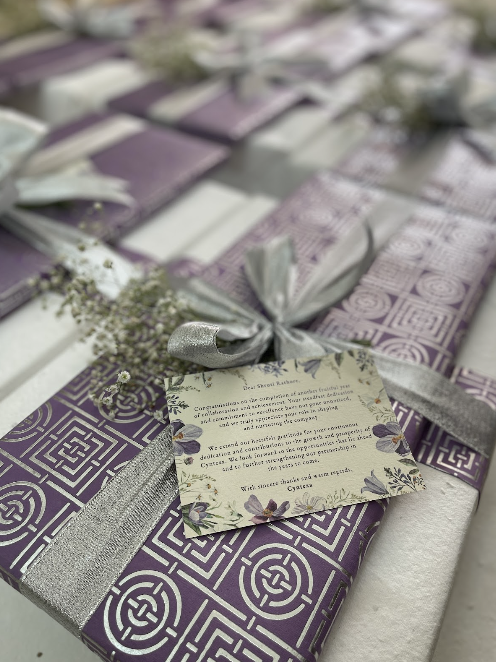 Purple and silver wrapped gift boxes with silver ribbons and small white flowers, featuring a floral card with a congratulatory message.