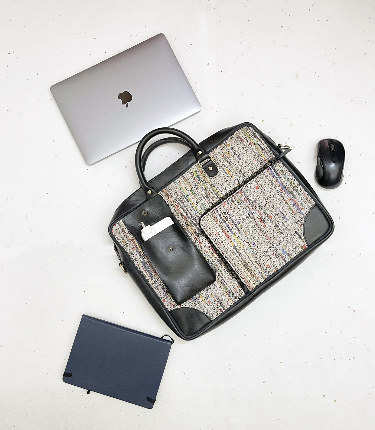 Flat lay of a patterned laptop bag with a white charger in side pocket, a closed silver MacBook, a black wireless mouse, and a closed navy notebook on a white surface.
