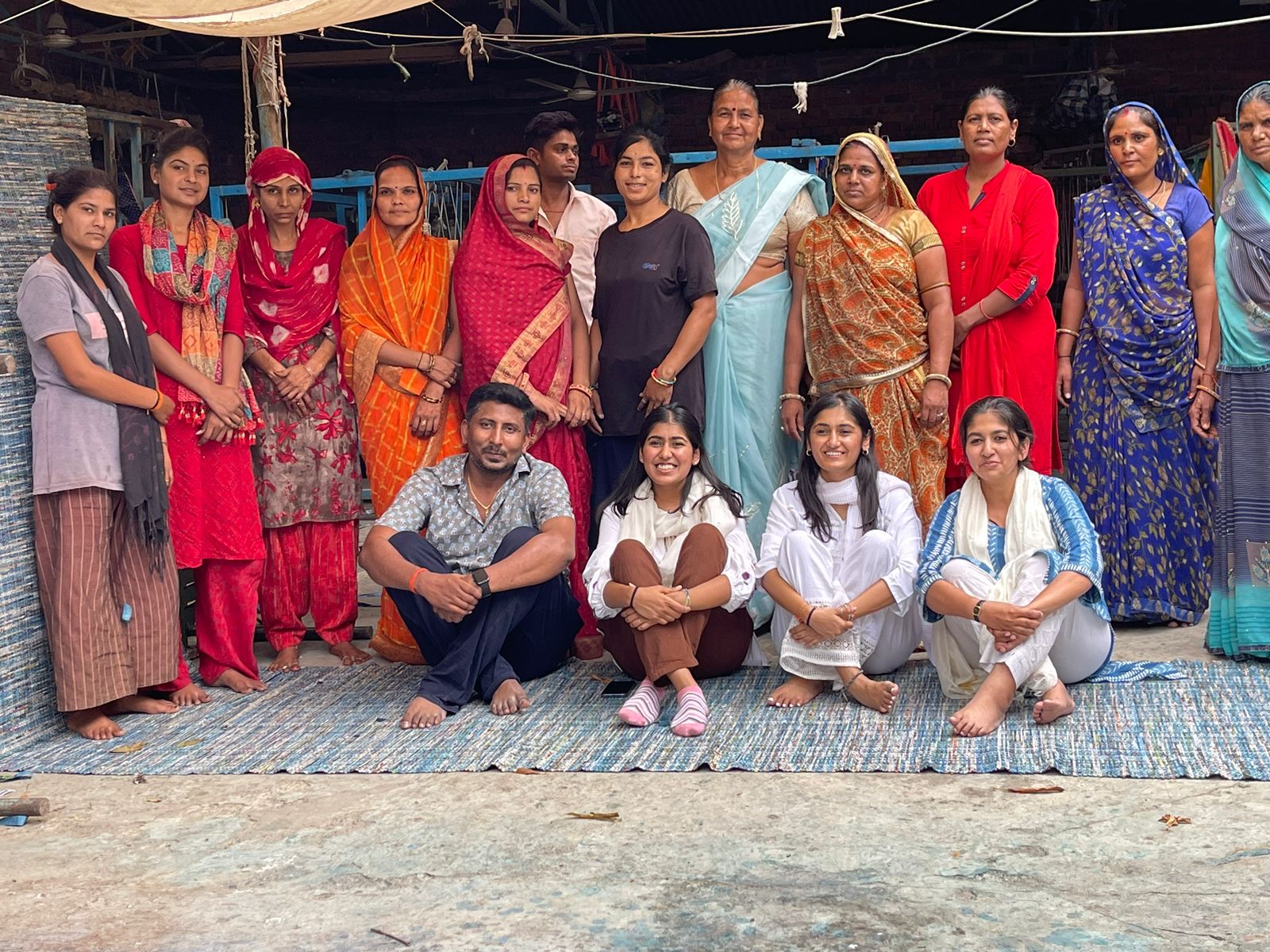 Group portrait of diverse men and women from India, some standing and some sitting on a woven mat in front of a rustic backdrop.