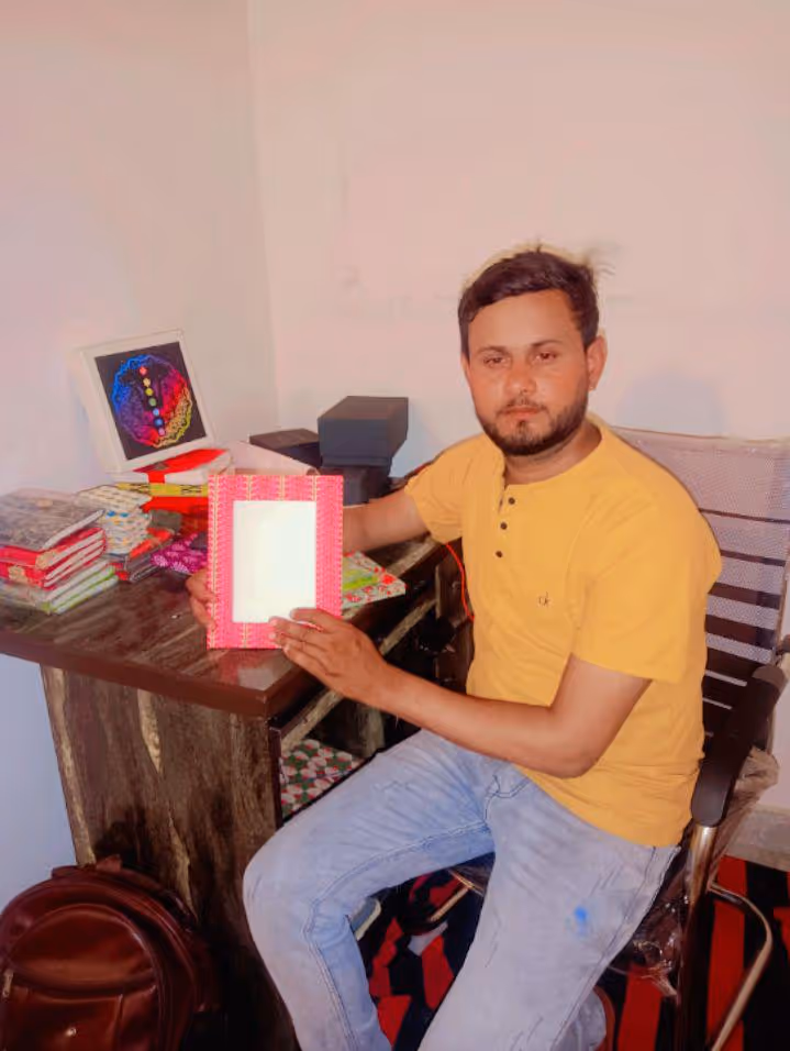 Man in a yellow shirt sitting at a desk holding a red patterned notebook with a blank white cover.
