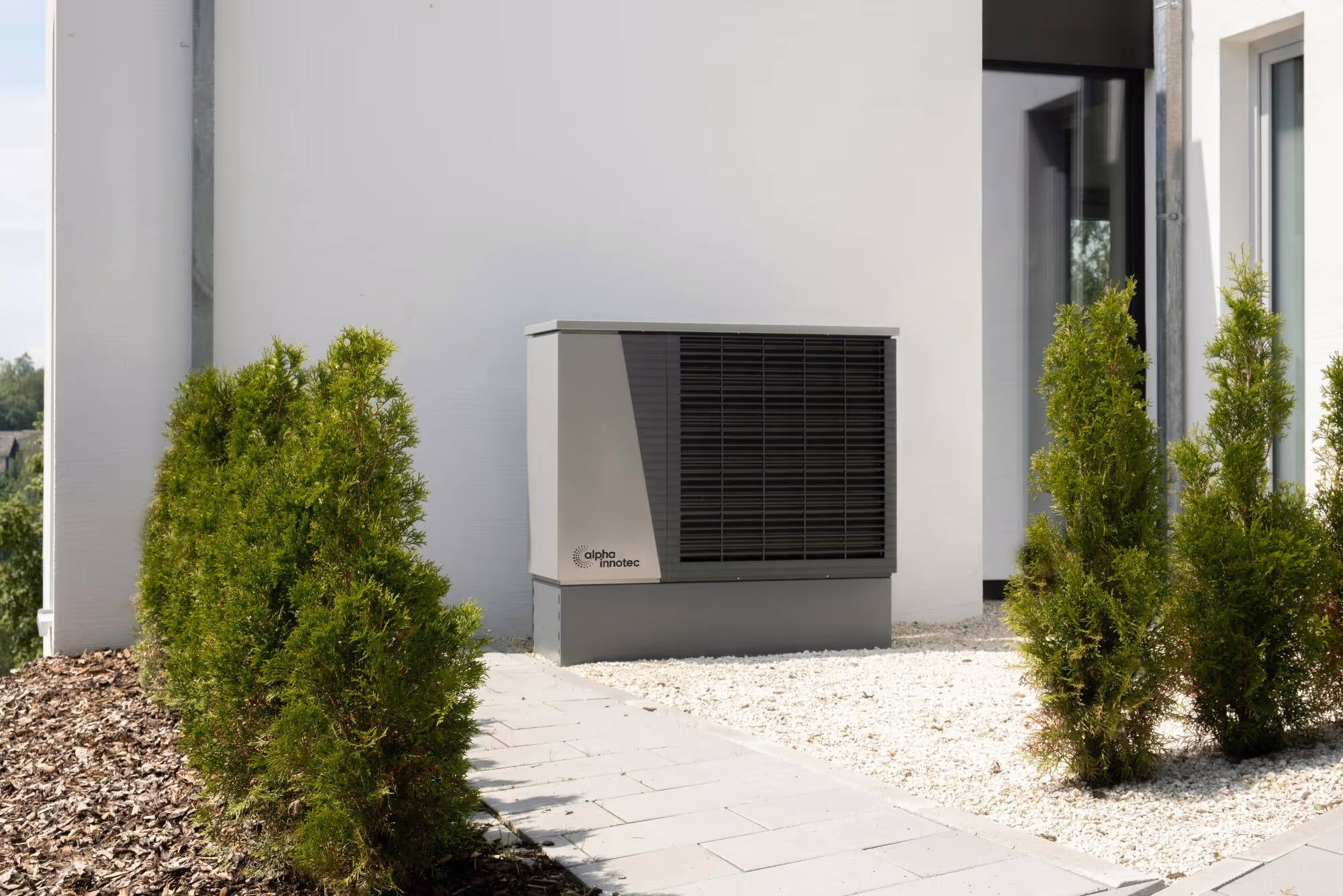 Alpha Innotec heat pump installed beside green shrubs on white patio