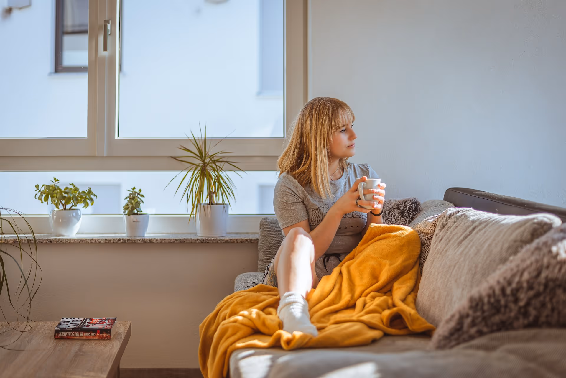 Person relaxing on couch with warm drink, yellow blanket, and indoor plants