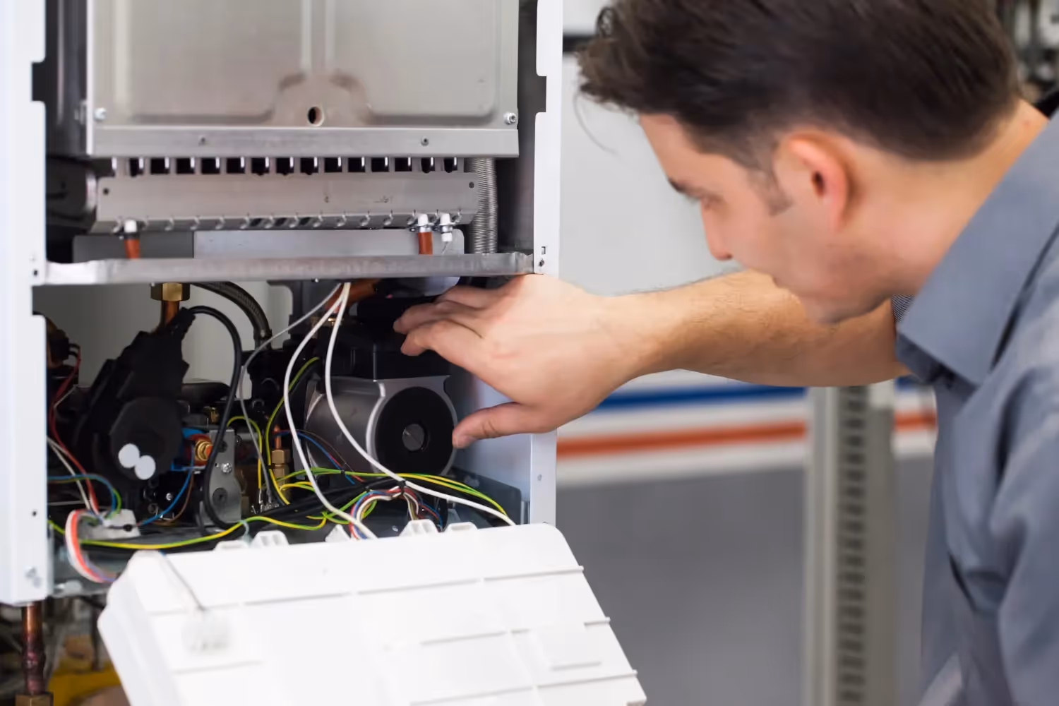 A person with dark hair is inspecting the internal components and wiring of a white furnace with its access panel partially removed.