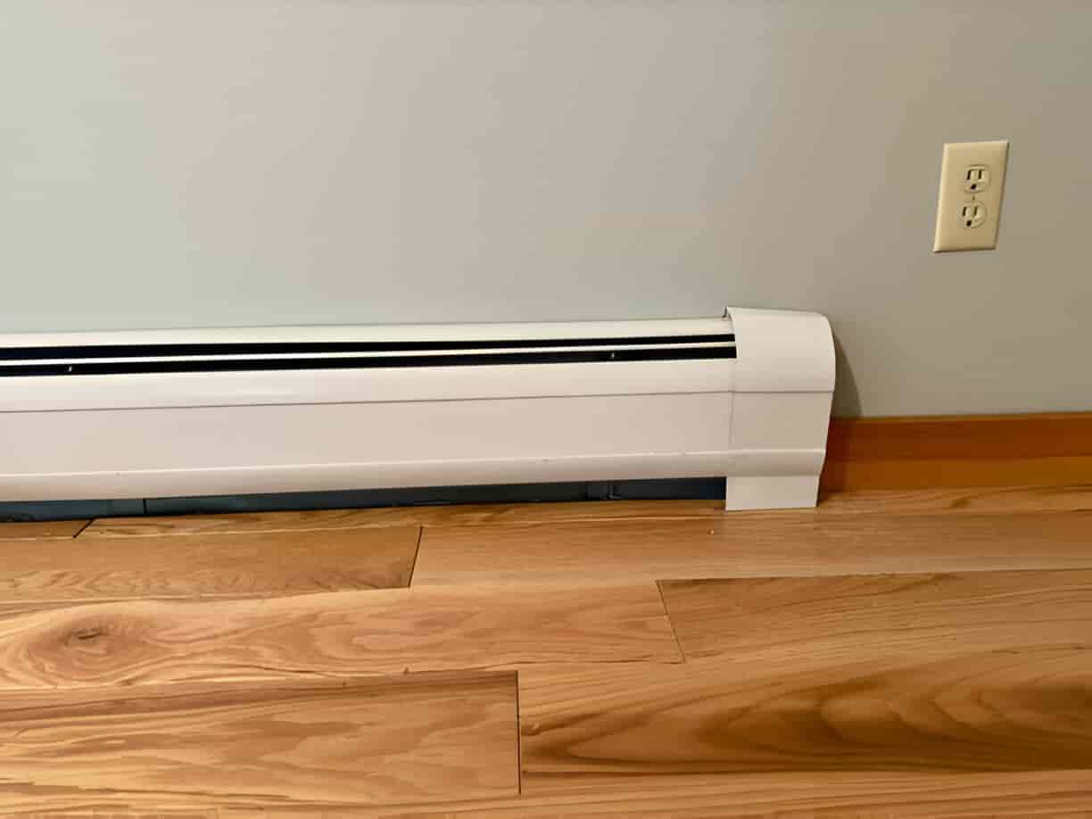 White electric baseboard heater mounted on wooden floor near wall outlet