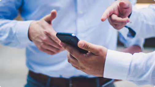 Two people in shirts using a smartphone, one pointing and the other holding the phone.