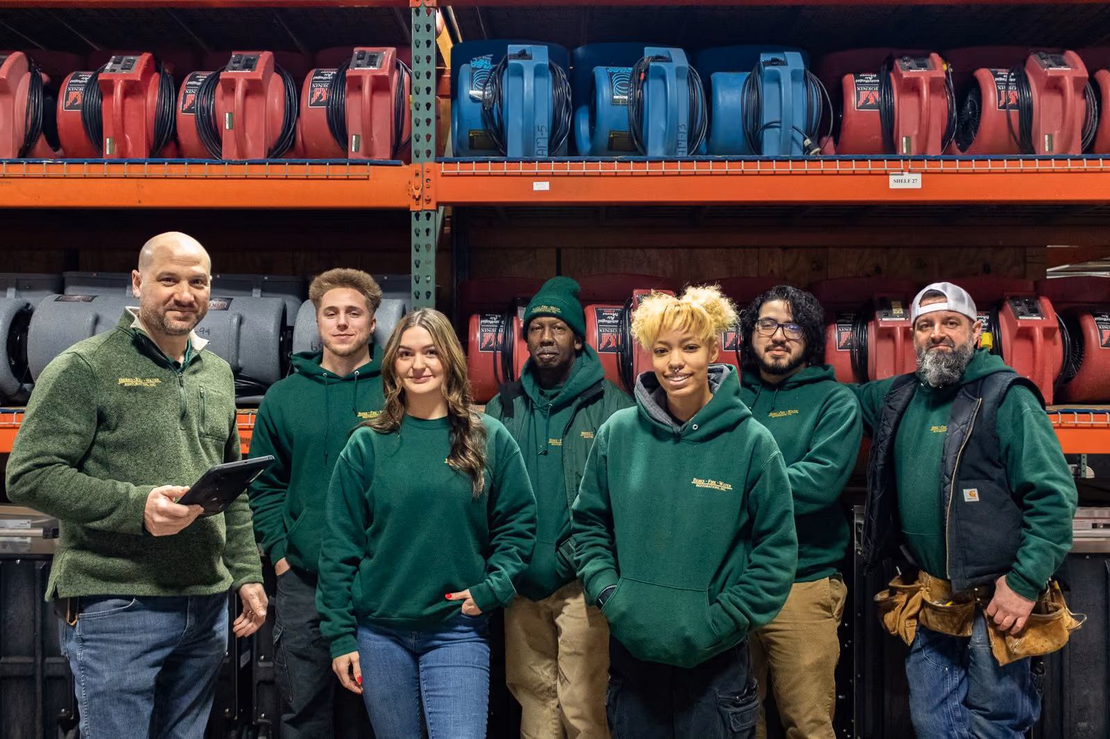 A restoration crew standing together in front of racks of air movers and drying equipment, with a manager holding a tablet.