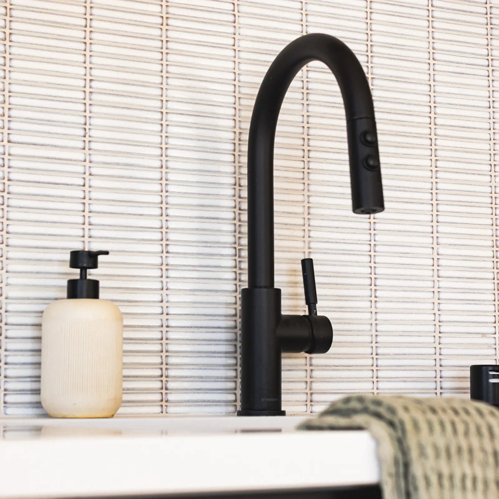 Matte black kitchen faucet with two buttons on the spout, next to a beige soap dispenser, against a white tiled wall.