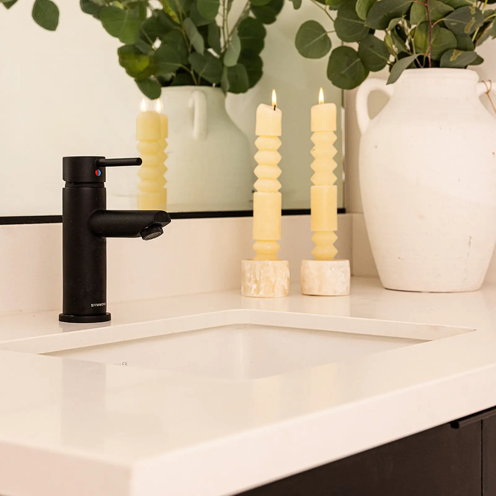White bathroom sink with black faucet, two lit yellow candles, and a white vase with green leafy branches in front of a mirror.