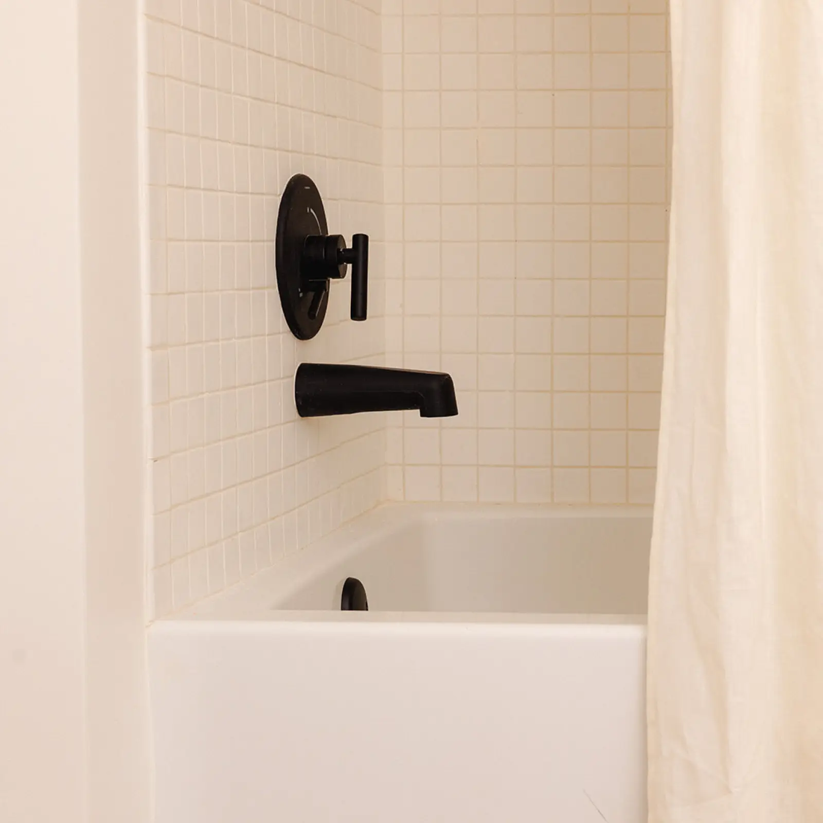 White bathtub with cream tiled walls and matte black faucet and handle, partially covered by a cream shower curtain.