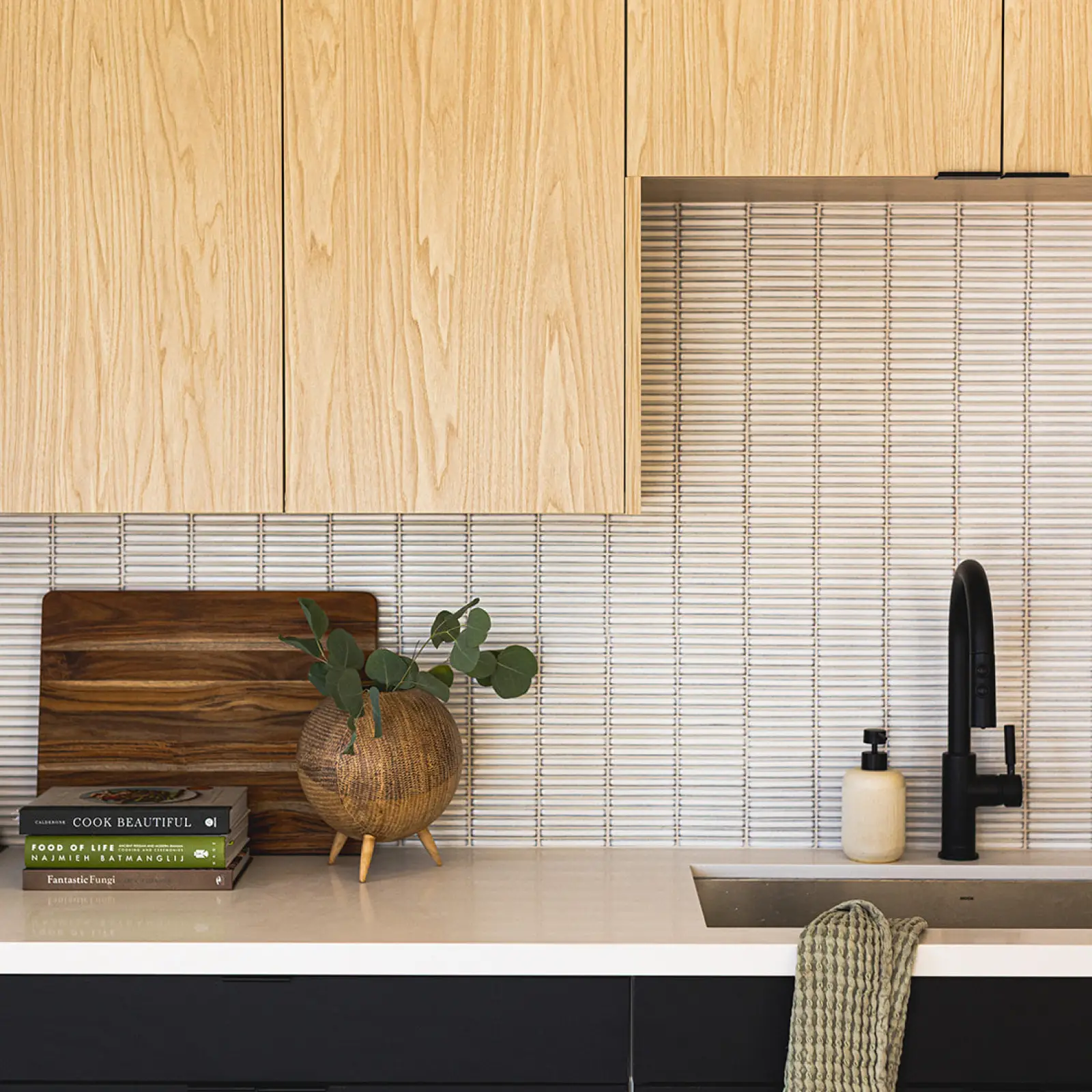 Modern kitchen countertop with wooden cabinets, a round wooden vase with greenery, cutting board, stacked cookbooks, white soap dispenser, black faucet, and a green towel hanging over the sink.