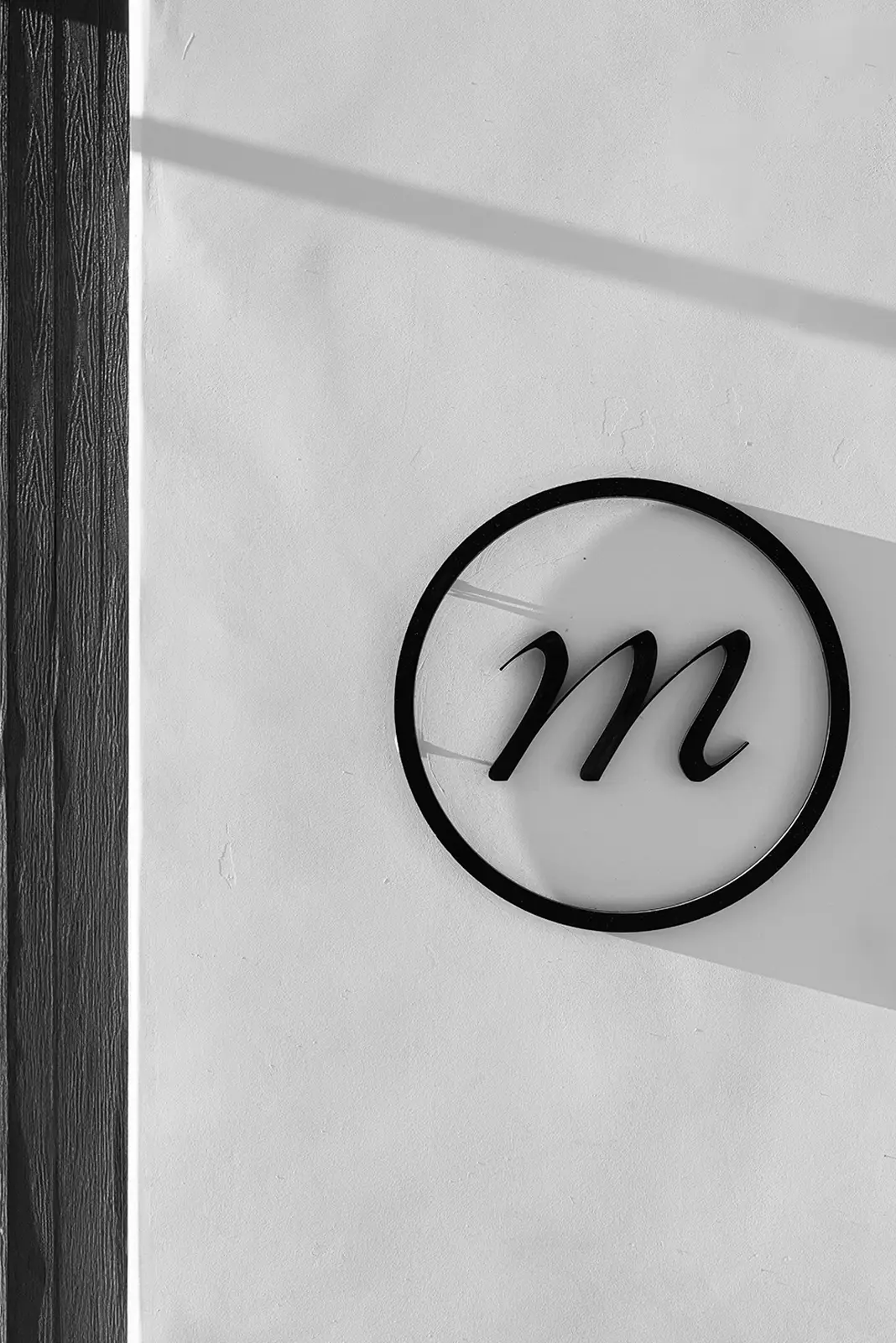 Mysa "m" mark on-building as a black metal circle with a lowercase letter 'm' mounted on a white textured wall casting a shadow.