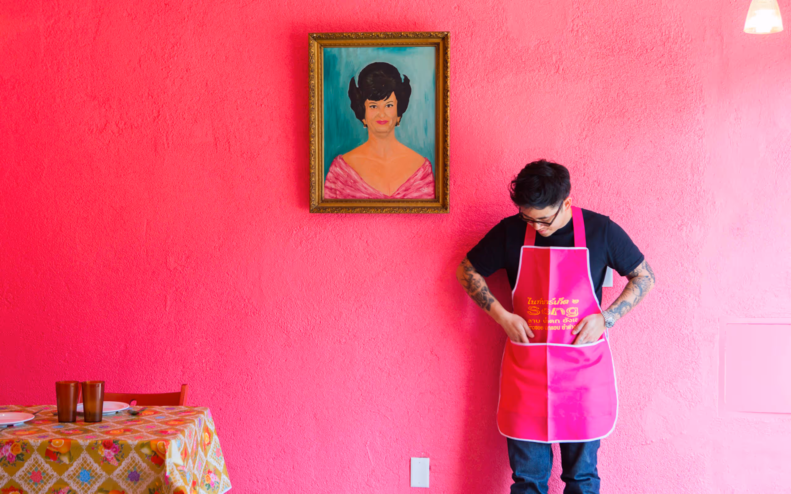 Person in a bright pink apron leaning on a bright pink wall with a painting of a women