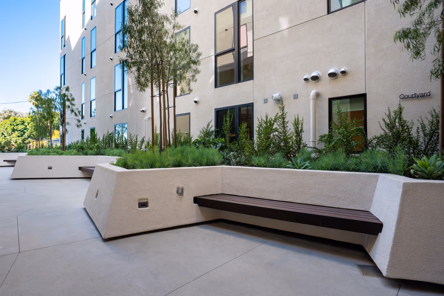 Modern apartment courtyard with geometric white planters, trees, shrubs, and beige building walls with windows.