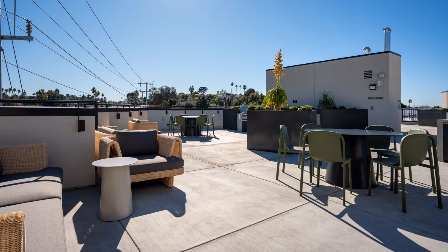 Sunny rooftop terrace with outdoor seating including green chairs around black tables and wicker armchairs with dark cushions.