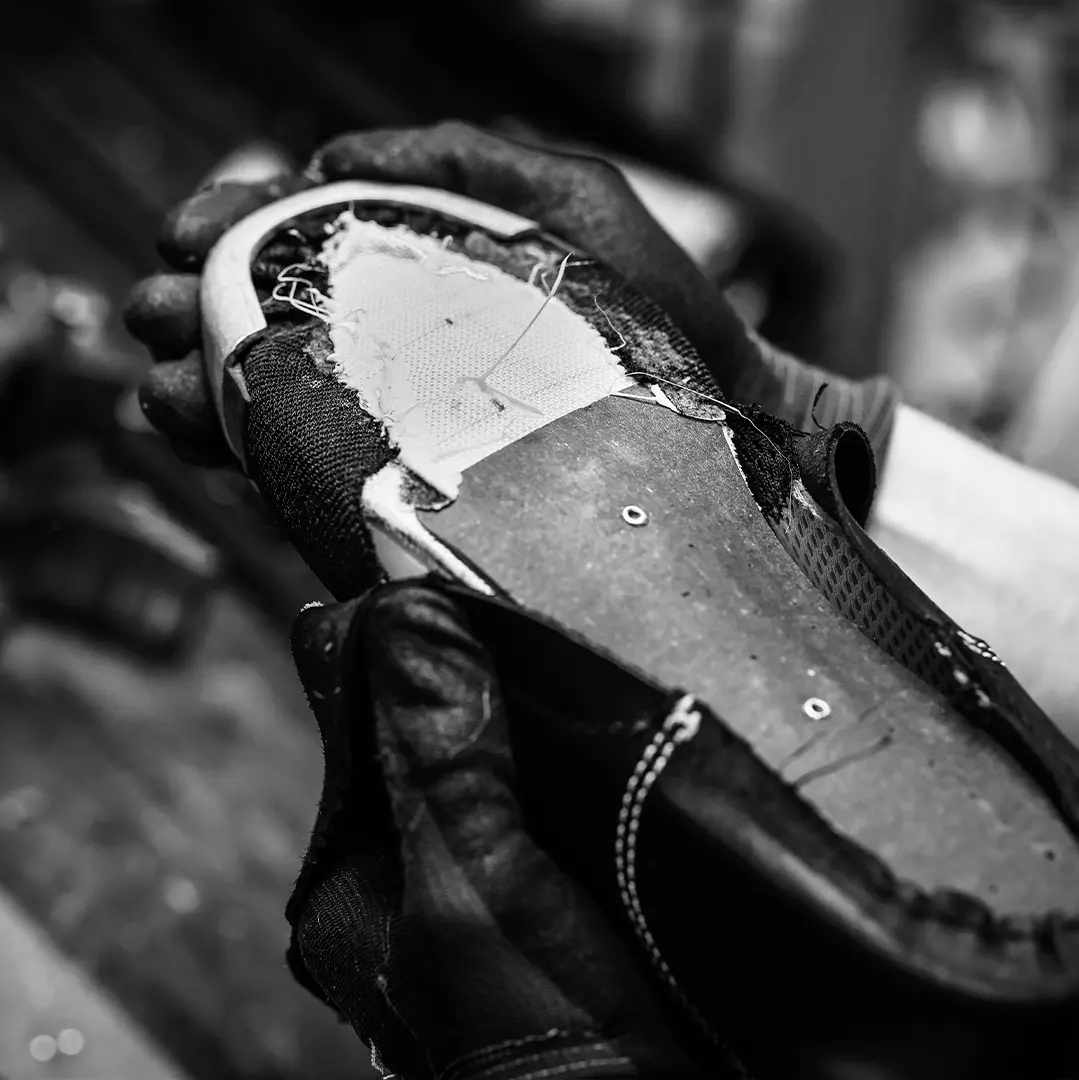 Svartvitt foto på händer med handskar som håller en sko under lagning med synliga sömmar och lappar.