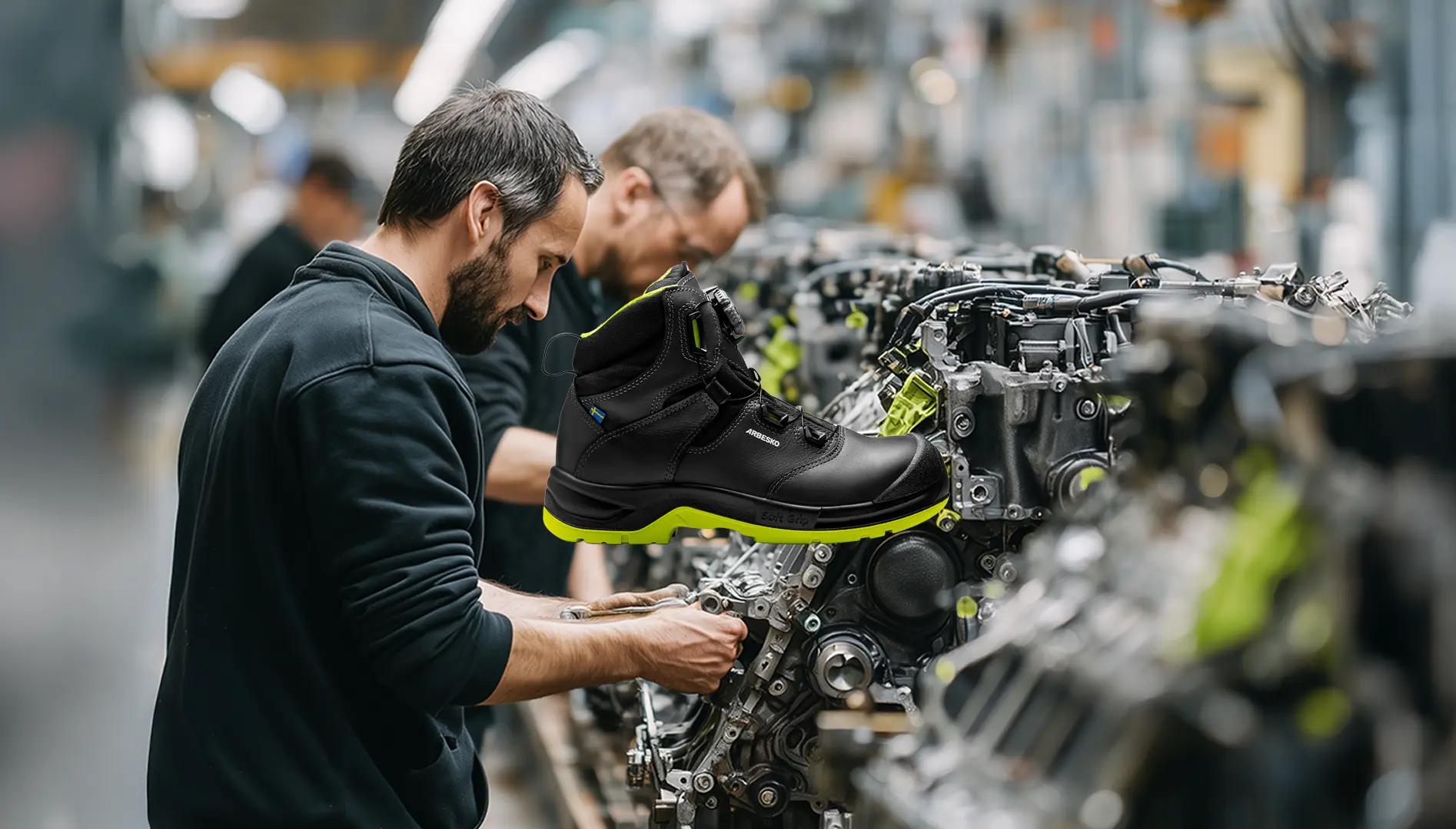 Två män som arbetar med motorer i en fabrik och en svart arbetskänga med gröna detaljer visas i förgrunden.