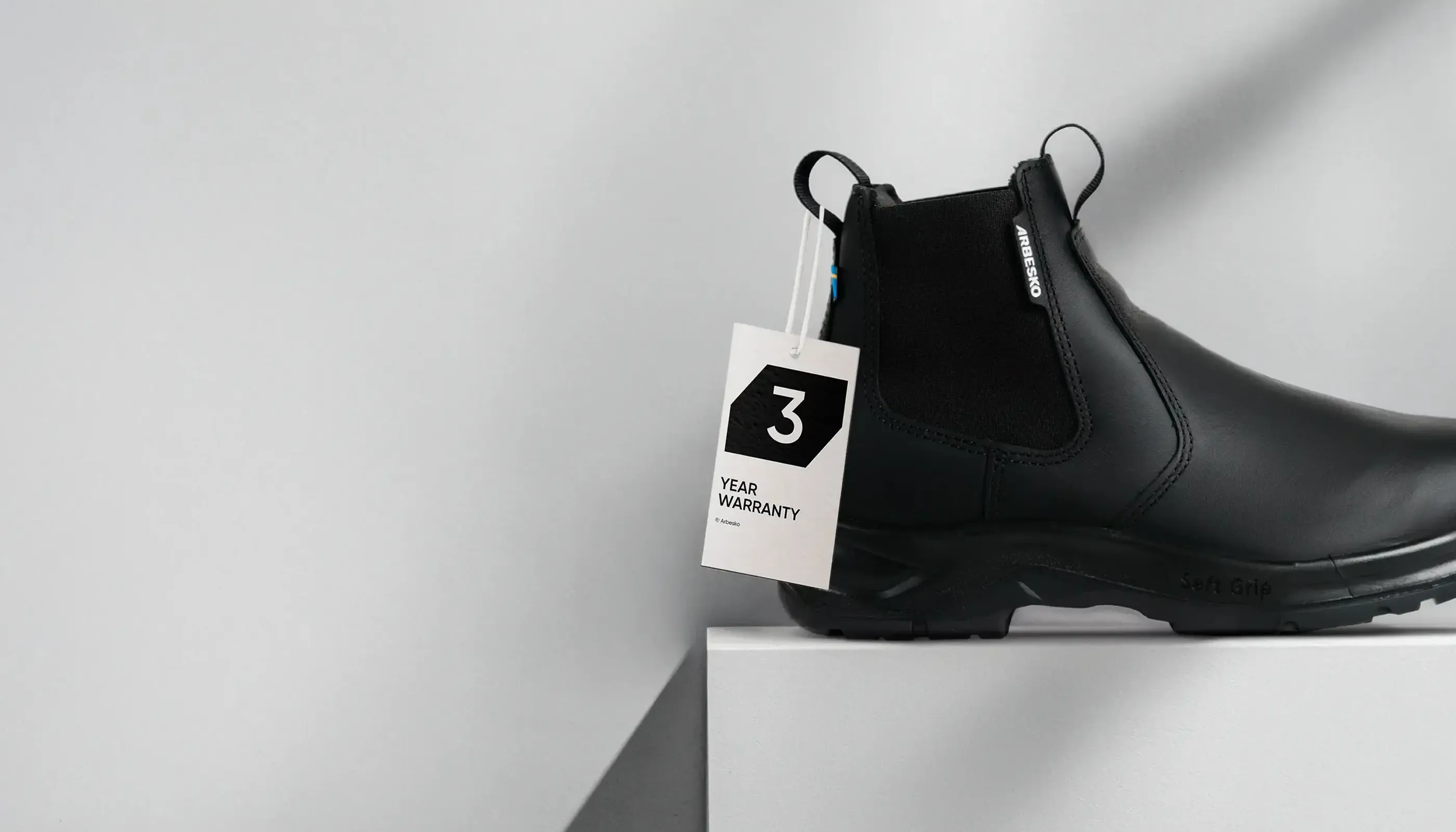 Svart Arbetsko med elastisk panel och etikett som visar 3 års garanti, placerad på vit piedestal.