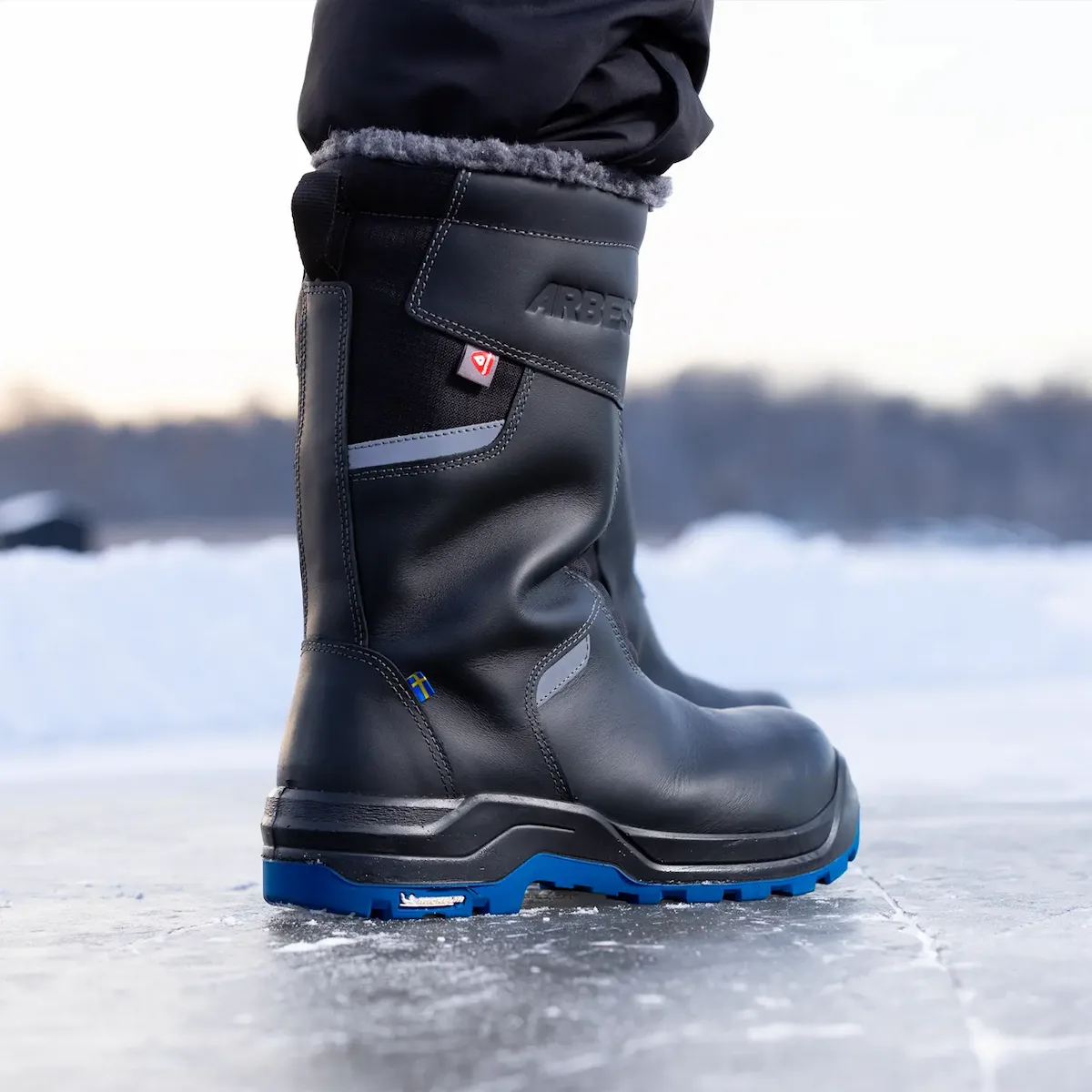 Närbild på svart vinterstövel med blå sula och sydd svensk flagga, stående på is.