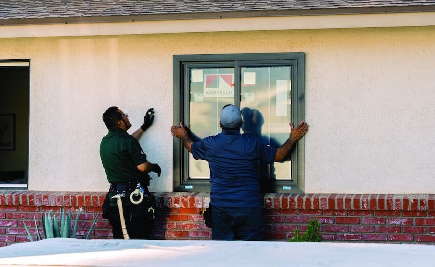 Two contractors coordinate an Andersen window from inside and outside an existing home opening during installation.