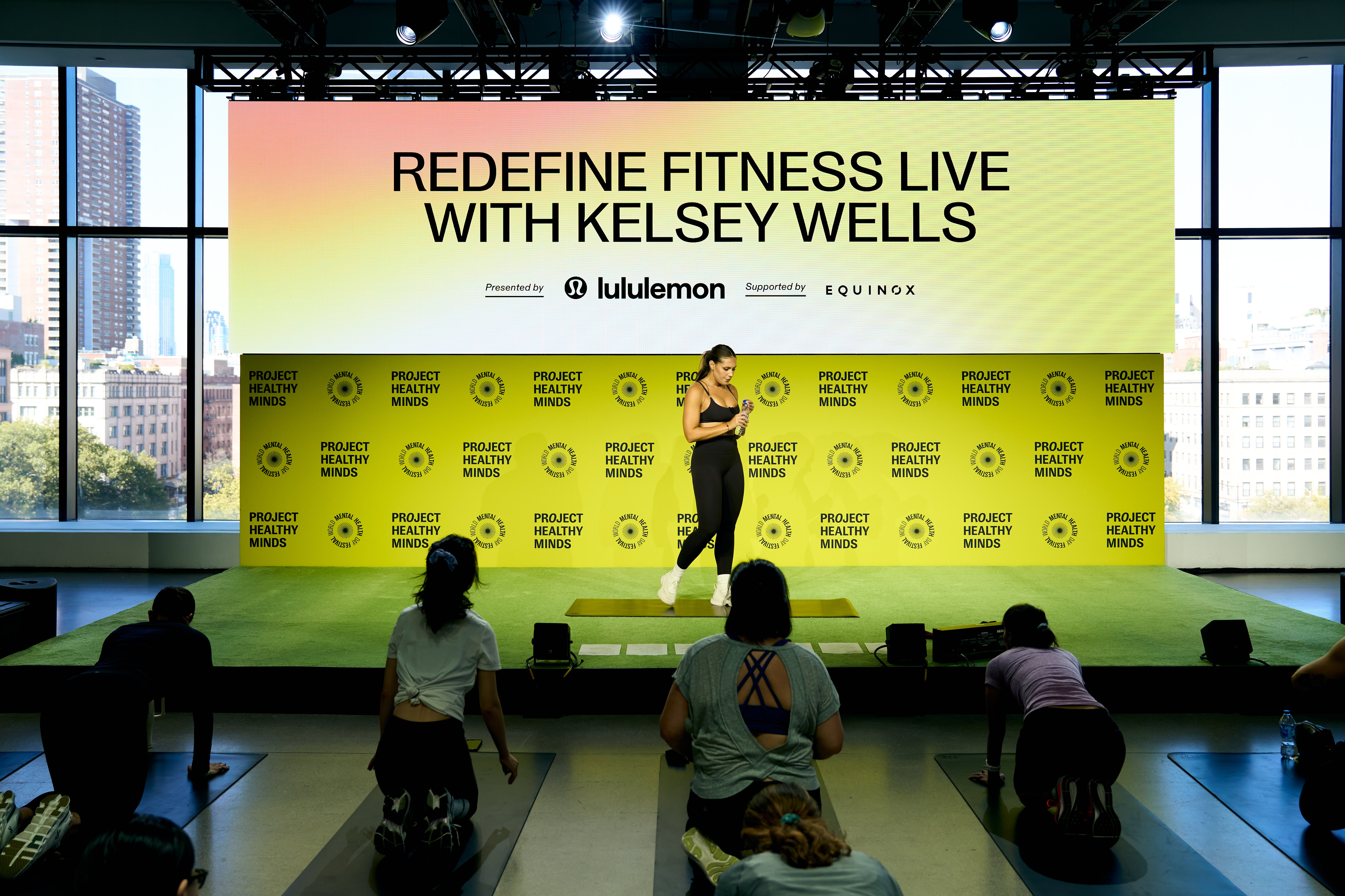 Fitness event with Kelsey Wells leading a workout on stage, Project Healthy Minds and lululemon branding behind her.