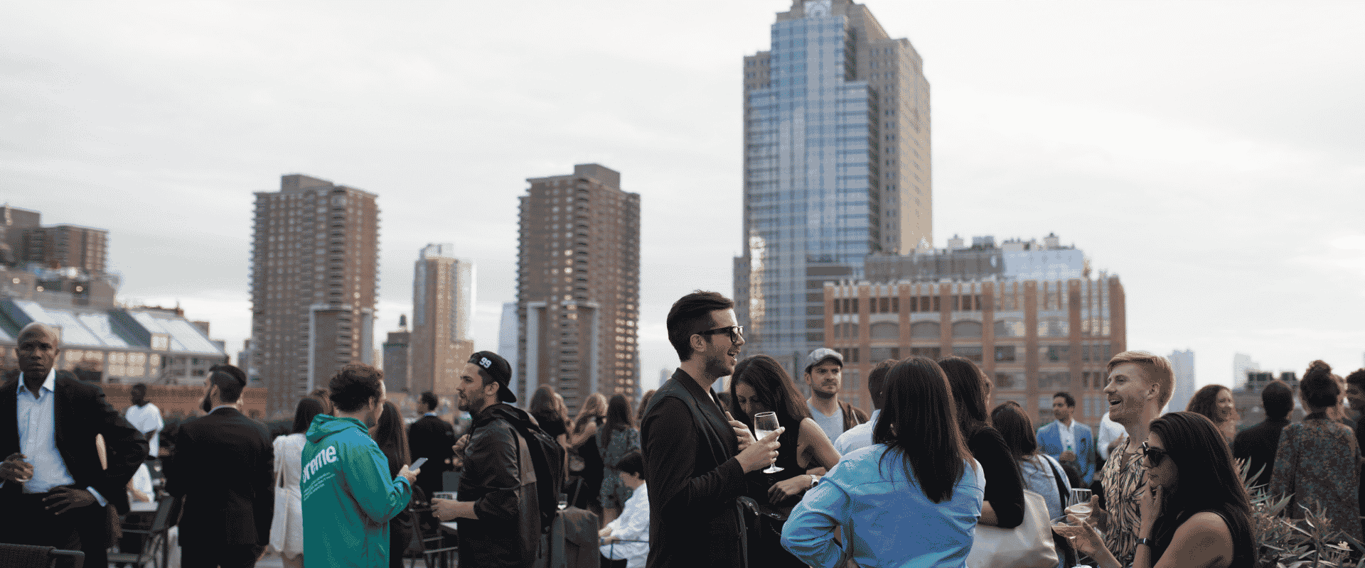 A crowd of people mingling at an outdoor event with a view of a city skyline.