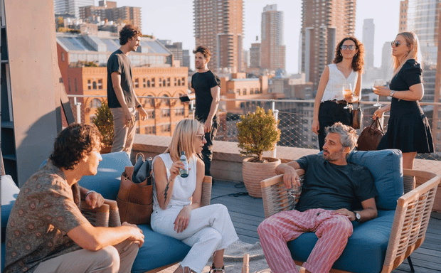 Group of friends socializing and enjoying drinks at a sunny rooftop event with city skyline views.