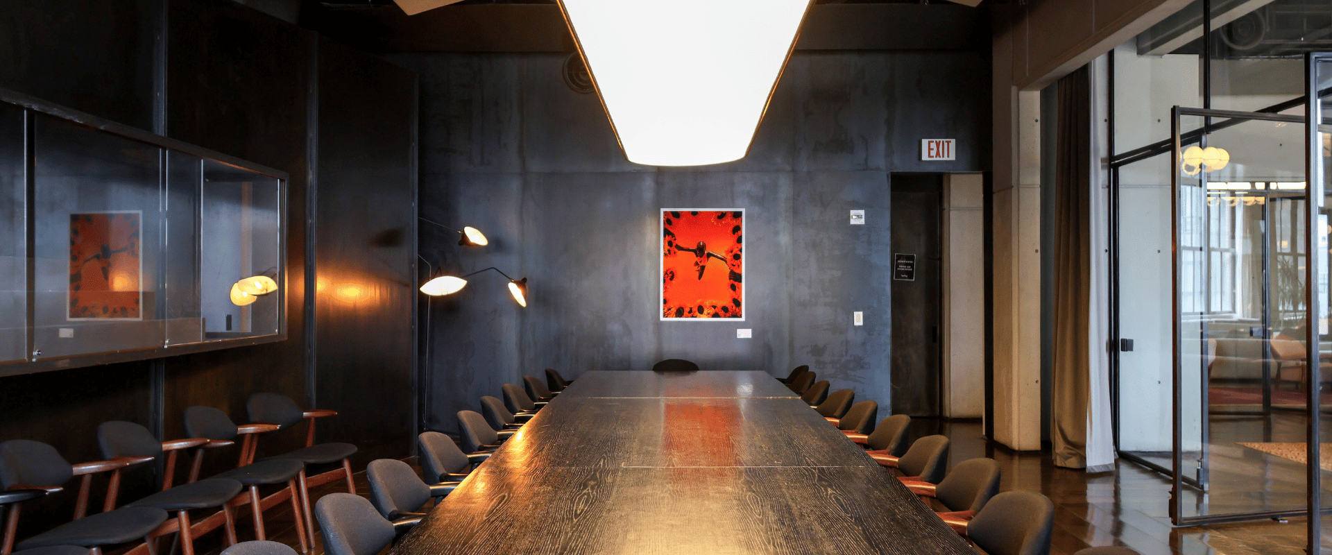 A sleek, modern conference room featuring a very long, dark wooden table lined with matching black chairs and illuminated by a massive, brightly lit pendant fixture.