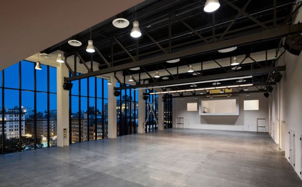 A spacious, empty event venue with industrial black ceilings, concrete floors, and expansive windows offering a twilight view of the city skyline.