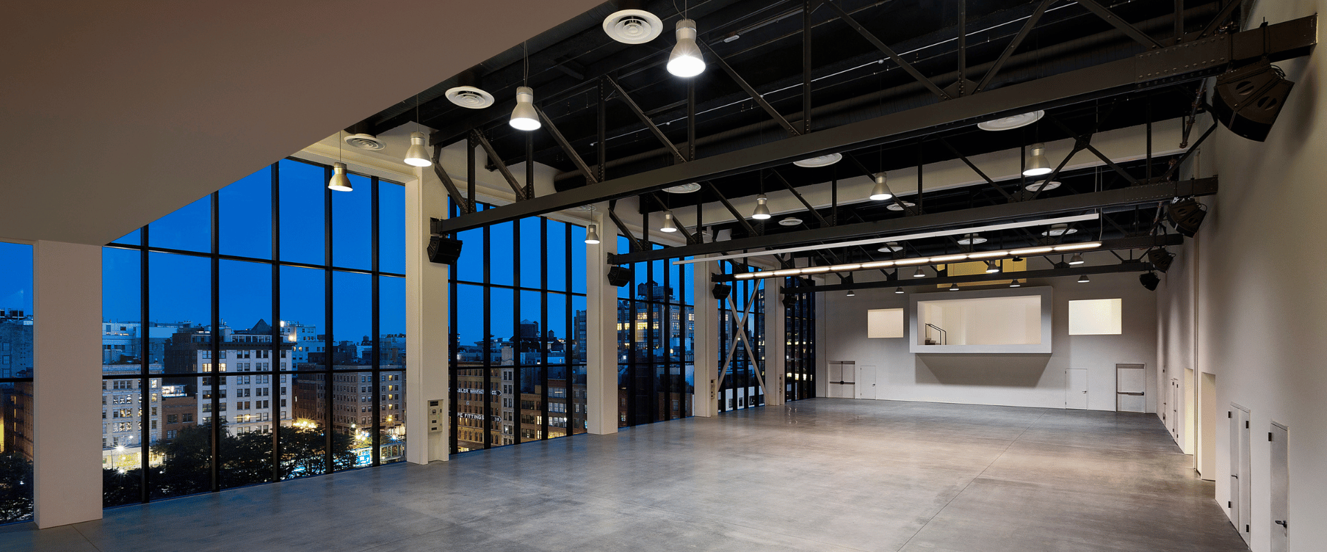 Large open-concept hall featuring floor-to-ceiling windows overlooking a city at dusk, highlighted by polished concrete floors and exposed steel ceiling trusses.