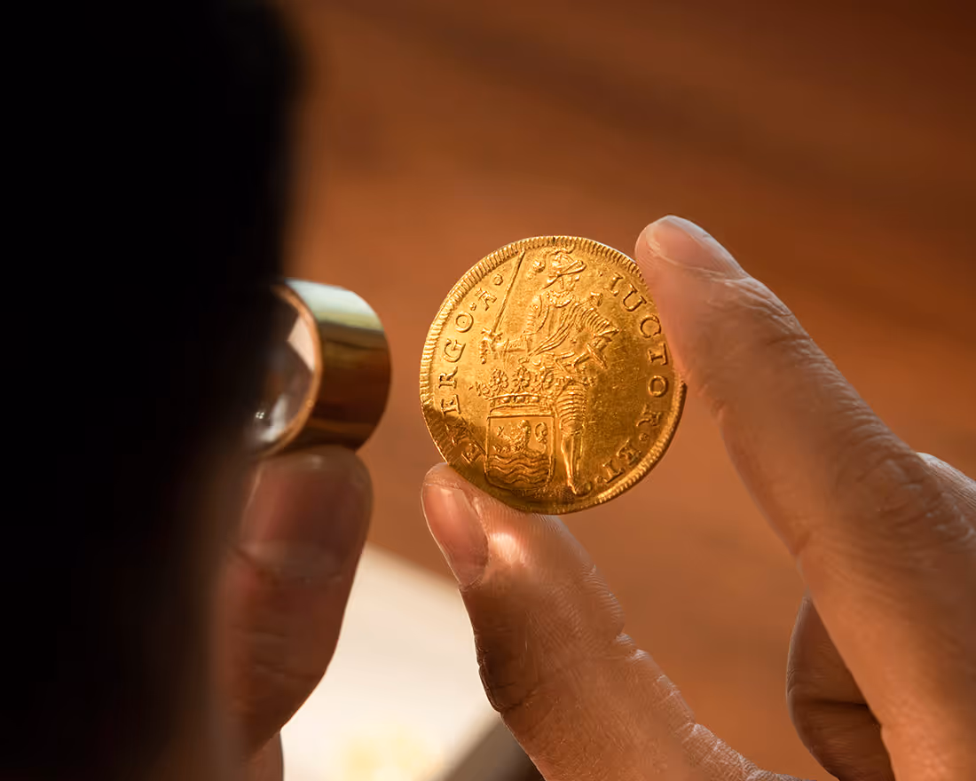 Hand inspecteert een gouden munt met een vergrootglas, met een afbeelding van een bewapende ridder en omringende tekst.