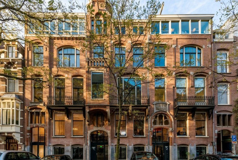 Vooraanzicht van een historisch kantoorgebouw met grote ramen, smeedijzeren balkons en een kale boom ervoor.
