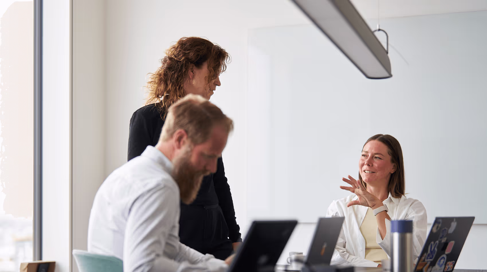 Bilden visar tre personer i en kontorsmiljö. En kvinna står och samtalar med en annan kvinna som sitter ner, medan en man med skägg och ljus skjorta arbetar vid en bärbar dator. Rummet har en ljus och modern atmosfär med en stor glasskiva på väggen.