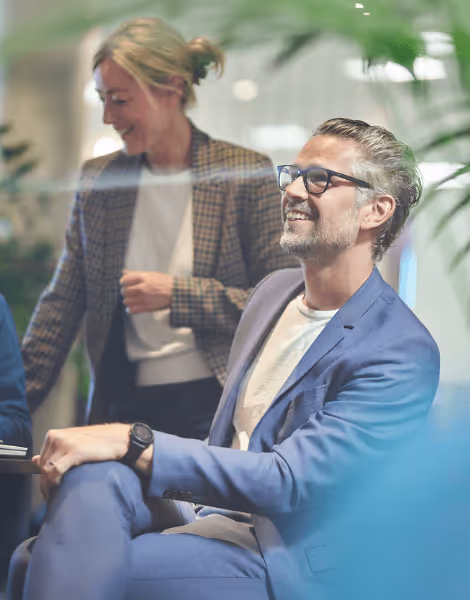 Bilden visar en leende man i blå kostym och glasögon som sitter ner i ett modernt kontorsområde. En kvinna i rutig blazer står bakom honom och ler, medan de omges av gröna växter.