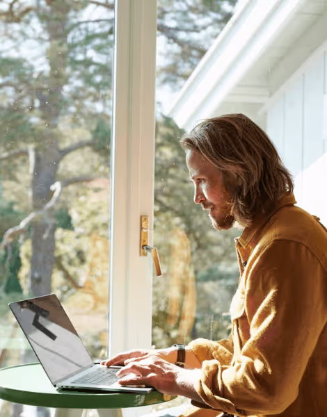 Bilden visar en man med långt hår och skägg som arbetar på en bärbar dator vid ett litet bord, medan solljuset strömmar in genom ett stort fönster. Han är klädd i en senapsgul skjorta och sitter i en miljö där naturen syns tydligt utanför.