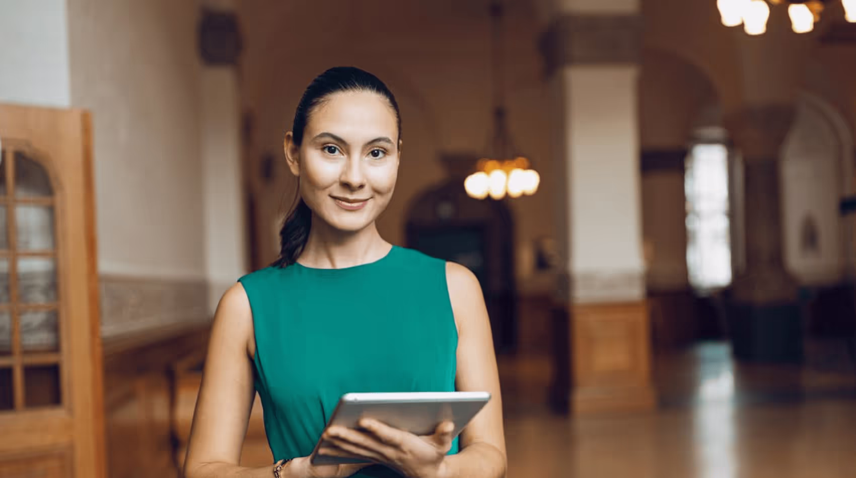 Bilden visar en kvinna med mörkt hår uppsatt i en hästsvans, klädd i en elegant grön klänning. Hon ler mildt och håller en surfplatta stående i en stor sal med varm belysning och arkitektoniska detaljer av trä och sten.