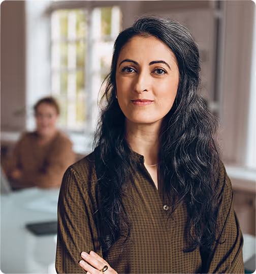 Bilden visar en kvinna med långt mörkt hår som står och ser självsäker ut mot kameran, klädd i en mörkbrun blus. I bakgrunden syns en annan person som sitter vid ett bord i ett rum med stora fönster och naturligt ljus.