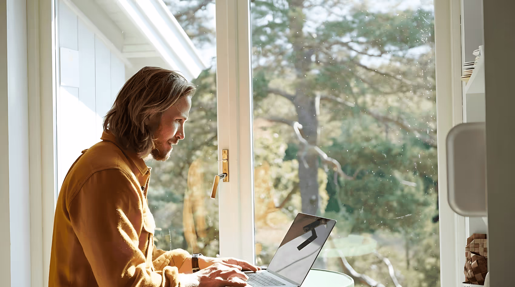 Bilden visar en man med långt hår och skägg som arbetar på en bärbar dator vid ett litet bord, medan solljuset strömmar in genom ett stort fönster. Han är klädd i en senapsgul skjorta och sitter i en miljö där naturen syns tydligt utanför.