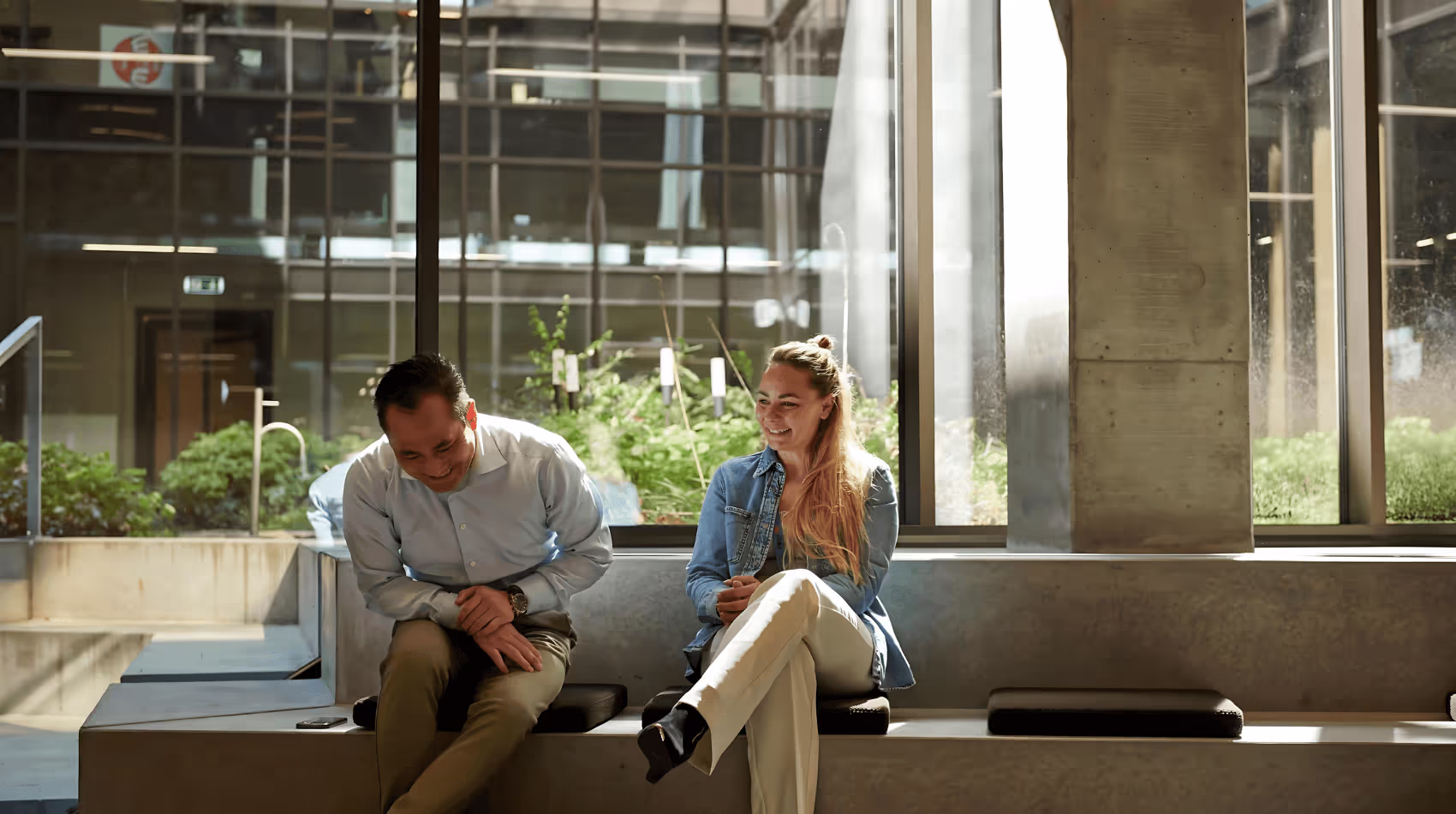 Bilden visar en man och en kvinna som sitter bredvid varandra på en modern bänk inomhus, med stora fönster som släpper in mycket ljus och ger utsikt över en grön innergård. Båda skrattar och verkar dela ett lättsamt ögonblick i en avslappnad arbetsmiljö.