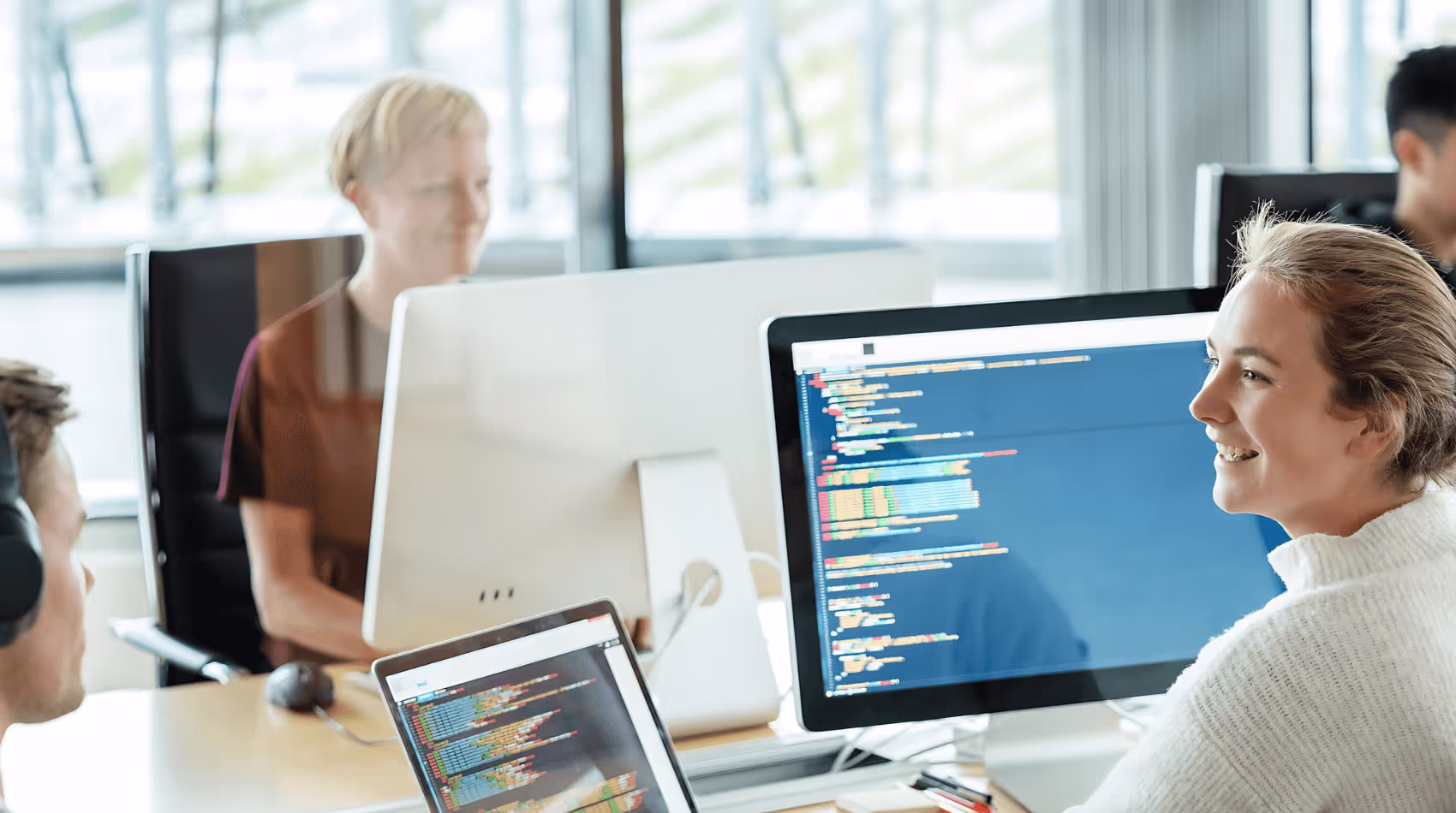 Bilden visar flera kollegor i en modern kontorsmiljö som arbetar vid sina datorer. På bildskärmarna syns kod, och ljuset strömmar in genom stora fönster, vilket skapar en ljus och produktiv arbetsatmosfär.