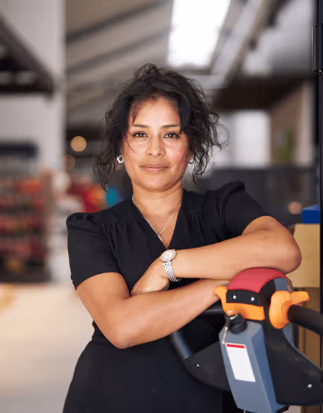 Bilden visar en kvinna med mörkt, lockigt hår som står med armarna korsade framför en palldragare i en lager- eller butiksomgivning. Hon är klädd i en svart blus och har en avslappnad och självsäker uppsyn.