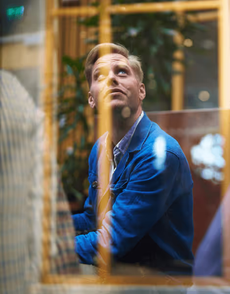 Bilden visar en man i blå kavaj som ser uppåt med ett tankfullt uttryck, fotograferad genom en glasvägg som skapar reflektioner. Bakom honom syns suddiga konturer av en kontorsmiljö med gröna växter.