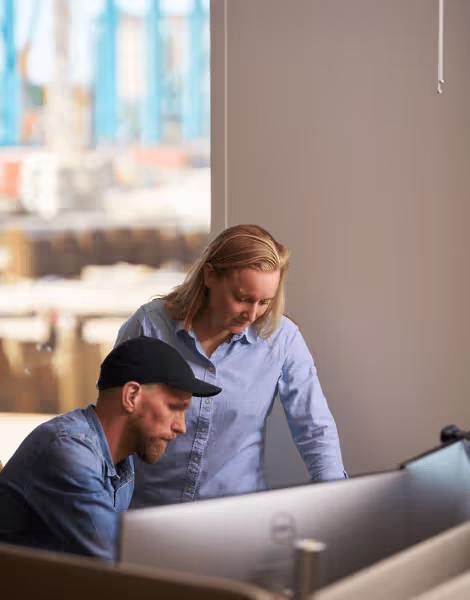 Bilden visar en man med keps och en kvinna, båda klädda i blå skjortor, som samarbetar framför en datorskärm i ett kontor. Genom fönstret ser man en byggarbetsplats med kranar i bakgrunden.