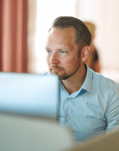 Bilden visar en man i ljusblå skjorta som ser fokuserad ut medan han arbetar vid en datorskärm i ett kontor. Bakom honom är bakgrunden suddig, med en annan person som delvis syns.