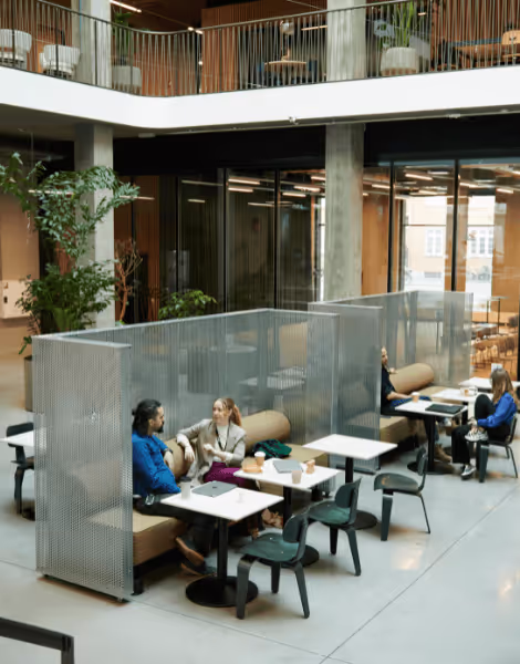 Bilden visar en modern kontorsmiljö med flera sittgrupper avskilda av skärmar för att skapa privata arbetsområden. Fyra personer sitter vid olika bord uppslukade av arbete och samtal, omgivna av växter och stora fönster som släpper in dagsljus.